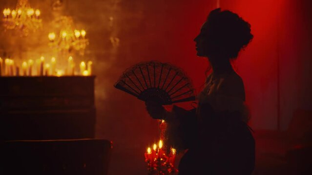 Art portrait dark shadow silhouette gothic sexy fantasy woman vampire queen holding in hands black fan in red light candles burning flame. Vintage dress Mystery noble lady girl night club room 4k