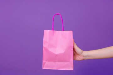 Womans hand holding pink paper shopping bag on purple background. Shopping sale delivery concept