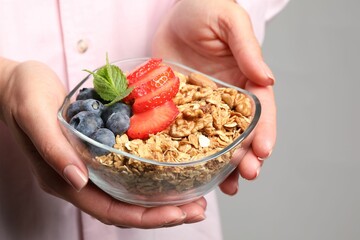 Woman holding bowl of tasty granola with berries, nuts and mint on light grey background, closeup