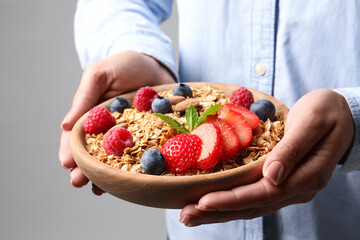 Woman holding bowl of tasty granola with berries, nuts and mint on light grey background, closeup