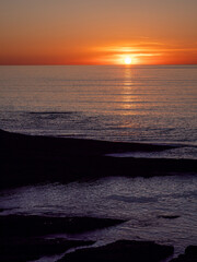 The sun is setting over the ocean, casting a warm glow on the water. The sky is a mix of orange and pink hues, creating a serene and peaceful atmosphere. The calm waters reflect the colors of the sky