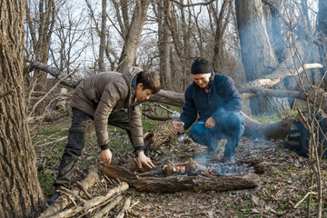 Two men make a campfire for cooking in the forest, father and son sitting on a log while hiking and outdoor activities, early spring landscape