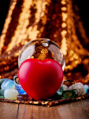 A heart is sitting on a table next to a crystal ball, golden textile with glitter. The scene is on wooden table, dark background. Still life image. Scene is romantic and mystical
