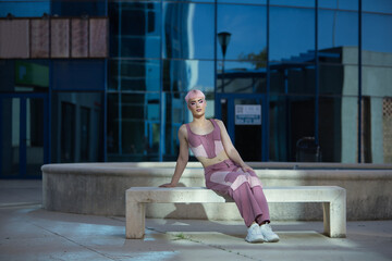 Fototapeta premium Young, attractive, gay man, heavily makeup, with pink hair, top and jeans, sitting on a stone bench, eyes closed, getting sun rays. Concept lgtbiq+, gay, pride, makeup, fashion, trend.