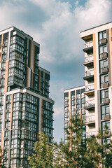 Fototapeta premium High-rise colorful residential buildings in a modern style against the sky with blue and white clouds. Houses of different geometric shapes with open balconies. Green trees grow in front.