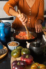 Closeup woman hands holding wooden spoon cooking, mixing chopped tomatoes stir fry in iron pan on stove. Homemade food concept