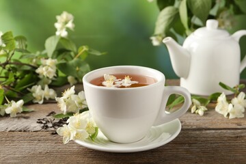 Aromatic jasmine tea in cup, flowers, green leaves and teapot on wooden table outdoors