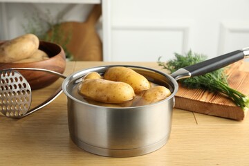 Raw potatoes in saucepan with water, pusher and dill on wooden table