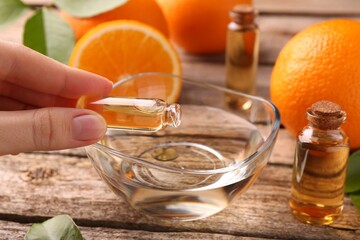 Woman pouring essential oil from cosmetic bottle into bowl at wooden table, closeup