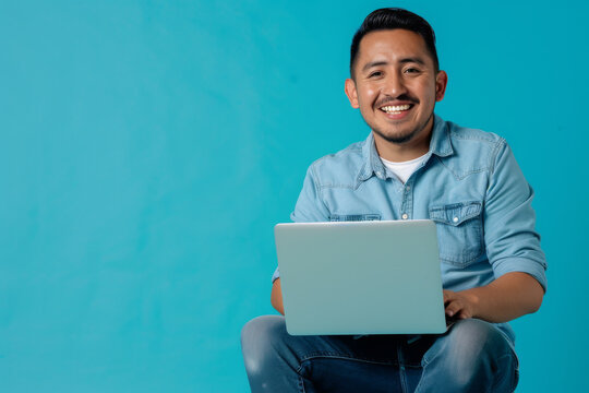 a Hispanic man in a blue casual shirt and jeans, looking happy while sitting with a laptop on a blue studio background