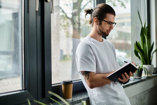 A man standing at a window, absorbed in reading a book.