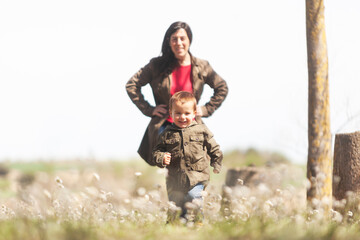 Fototapeta premium Toodler running through a meadow full of flowers. His mother watches him from behind.