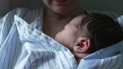 Close-up of a newborn baby sleeping on the mother's chest, wrapped in a blue and white striped shirt. The image highlights the tender moment of skin-to-skin contact between mother and baby