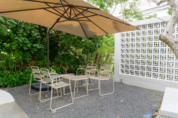 An outdoor seating area featuring beige chairs and a large umbrella, surrounded by lush greenery and a white decorative wall, providing a shaded spot for relaxation.