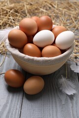 Fresh chicken eggs in bowl, feathers and dried straw on grey wooden table