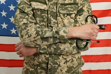 Fototapeta premium Man in military uniform with crossed arms and stethoscope against USA flag, closeup. Health care concept