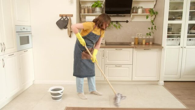 A woman mops the floor in her home kitchen. Female hands in yellow gloves while cleaning the kitchen