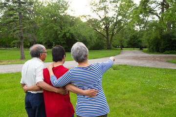 Three people are standing in a grassy field, with a woman pointing to the right. The man and woman are older, and the young man is wearing a red shirt. Scene is peaceful and relaxed