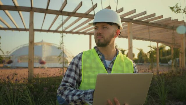 Thoughtful Caucasian male man experienced worker inspector helmet hardhat guy engineer technician typing laptop computer entering data internet pc analyzing project working blueprint construction park
