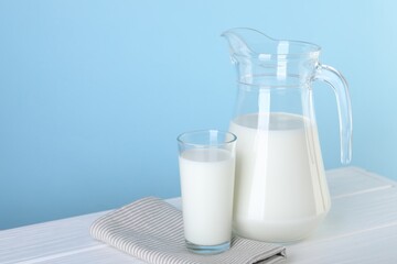 Jug and glass of fresh milk on white wooden table against light blue background, space for text