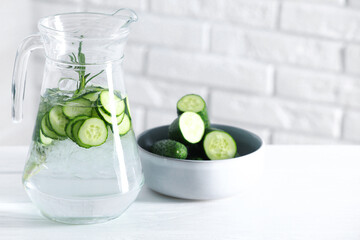 Refreshing cucumber water with rosemary in jug and vegetables on white table against brick wall, closeup. Space for text