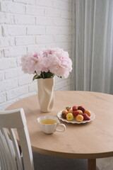 Ceramic wase with flowers, cup of green tea and plate with fruits and berries on wooden round table in light kitchen.