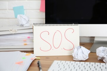 Notebook with word SOS, computer and stationery on wooden table at workplace