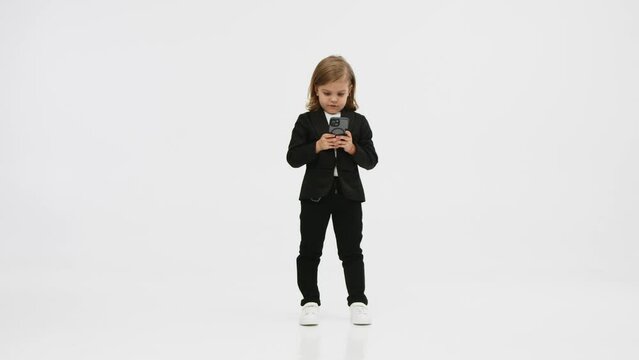 Little business girl on a white background in the studio clicks on a mobile phone and shows a "super" gesture of approval. Concept of business, deal and modern technology.