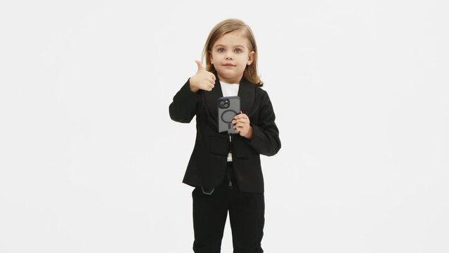 Little business girl on a white background in the studio clicks on a mobile phone and shows a "super" gesture of approval. Concept of business, deal and modern technology.