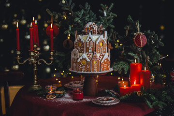 Elegant luxury Christmas table setting for family home party with homemade big gingerbread house, holiday celebration. Red tablecloth, candles, chandelier. Romantic candlelit dinner