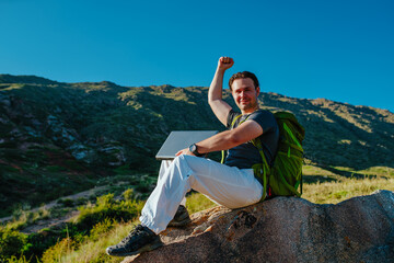 Young successful tourist with backpack and laptop sits on big stone on mountains background