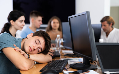 Exhausted young employee engaged in software development at IT startup in coworking space sleeping at workplace due to intensive work..
