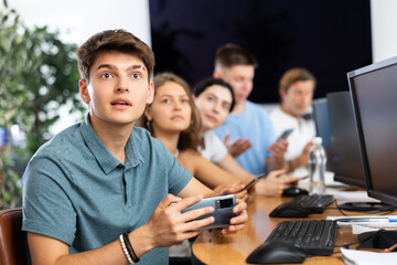 Positive guy student of computer courses sits among students and holds mobile phone in hands, plays game, tests new application