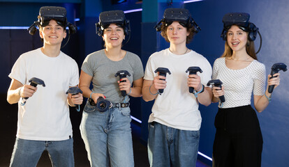 Group of cheerful young people, girls and guys, with handheld controllers in hands and VR glasses...