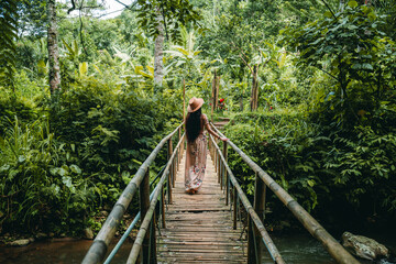 The young woman in a dress and hat is walking on the bridge, looking around in a tropical forest. Leke Leke Waterfalll Trail. Travel concept.