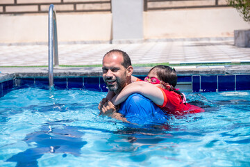 A young father supports his daughter in the swimming pool, helping her learn to swim and enjoying a wonderful time together playing in the water