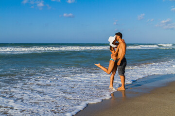 Happy young couple in bikini and shorts enjoying summer dusk at the beach, having fun walking...