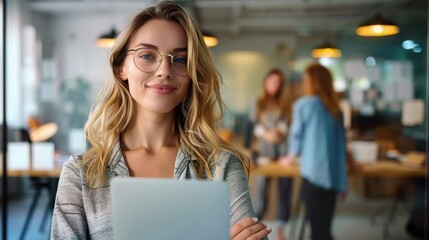 A professional woman with glasses smiles while holding a laptop, standing in an office environment