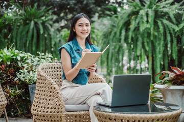 A woman is sitting on a chair with a laptop and a notebook in front of her. She is smiling and she is enjoying her work. The scene suggests a relaxed and comfortable atmosphere