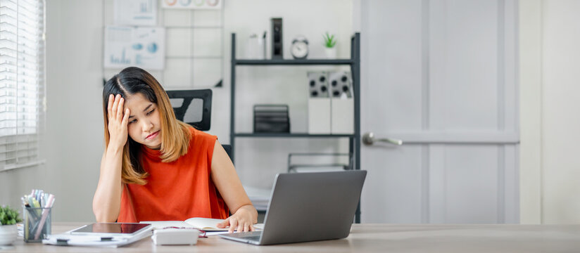 A woman in an orange shirt is sitting at a desk with a laptop and a stack of papers. She is in a state of distress or frustration