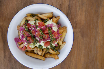 Top view of gourmet fried potatoes with avocado cream, red onion, tomato and pesto, in a white bowl on the wooden table. 