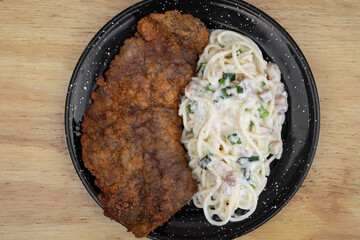 Pasta and meat. Top view of a milanesa, breaded steak, and spaghetti with a bacon and leek cream sauce, in a black dish on the restaurant wooden table.