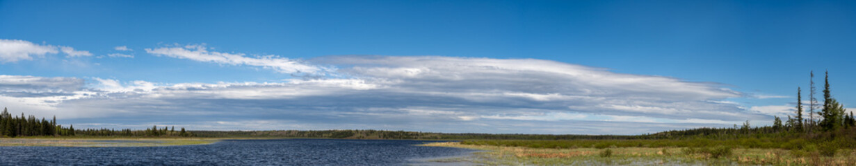 A wide panoramic view of a small lake surrounded by a forest. The blue sky is partially covered in a large white to gray cloud.
