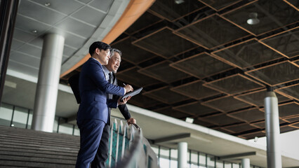 Fototapeta premium Two men in suits stood on the stairs. One of them is holding a tablet. The scene is professional and serious. This is because men often discuss important business matters.