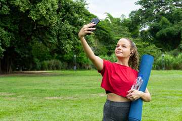 Young hispanic woman taking selfie communicating with smartphone holding yoga mat and water bottle in a park. Using earbuds. Exercise and workout.