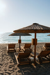 Sunny beach landscape with wooden sun loungers and straw umbrellas on sandy shore facing the sea.