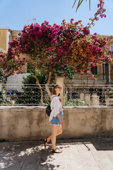 A woman strolls down a quaint street, passing by a vibrantly blooming tree under the clear, sunny...