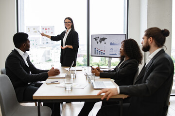 Business woman delivering presentation to diverse colleagues. Team meeting with graphs and charts near panoramic window. Professional setting with laptops and documents.