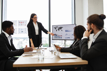 Business woman gives report to multiracial colleagues standing near graphs on monitor. Team meeting with analytics and performance charts. Professional setting with diverse team.