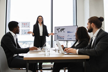 Business woman gives report to multiracial colleagues standing near graphs on monitor, screen, display, TV. Professional team in formal attire discussing data analysis, financial growth, statistics